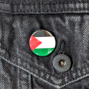 May include: A round button pin featuring the flag of Palestine, with red, white, green, and black colors. The pin is attached to a black denim jacket. A silver button is visible.