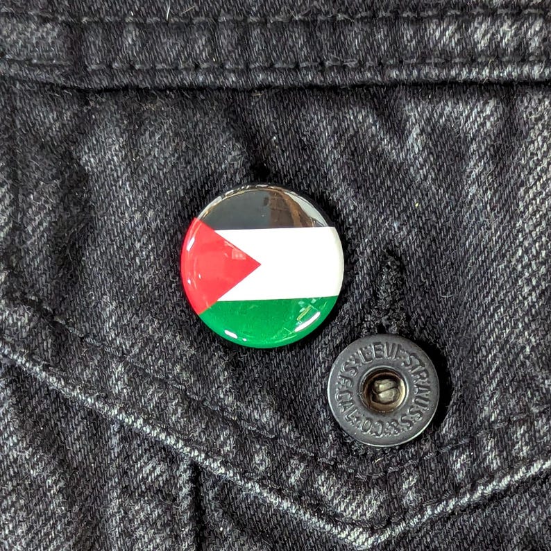 May include: A round button pin featuring the flag of Palestine, with red, white, green, and black colors. The pin is attached to a black denim jacket. A silver button is visible.