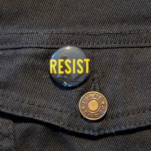 May include: A black button with the word "RESIST" in yellow lettering on a black denim jacket.