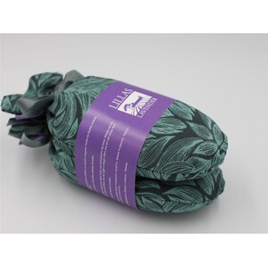 Puede incluir: Un saquito de lavanda con un estampado de hojas verde azulado y negro. El saquito está envuelto con una etiqueta morada que dice "LILLAS LAVENDER". El saquito está atado con una cinta gris.