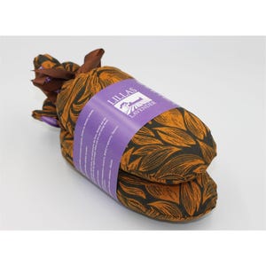 Op de afbeelding: Twee met lavendel gevulde kussens, verpakt in stof met een bladpatroon in oranje en zwart. Een paars etiket met de tekst "LILLAS LAVENDER" is om de kussens gewikkeld. De kussens zijn vastgebonden met een bruin lint.