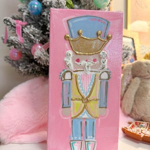 May include: A pink canvas painting of a nutcracker soldier. The nutcracker has a blue hat, gold crown, and a yellow vest. The painting is decorated with pink, blue, gold, and white paint. The painting is displayed in front of a small Christmas tree.
