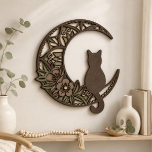 May include: A wooden wall art piece featuring a cat silhouette perched on a crescent moon. The moon is intricately designed with floral and geometric patterns in shades of brown, green, and beige. The cat is a solid brown color.