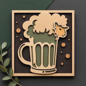 Può includere: Opera d'arte da parete in legno raffigurante un boccale di birra con la testa di una pecora che spunta dalla schiuma. Il design utilizza tonalità verdi e marroni a strati, su uno sfondo scuro con cerchi di legno. L'opera è incorniciata in legno marrone chiaro.