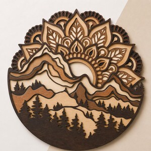 May include: A circular multi-layered wooden wall art piece featuring a mountain landscape with a forest of pine trees in the foreground. The design incorporates a sunburst mandala pattern in shades of dark brown, medium brown, and light beige wood.