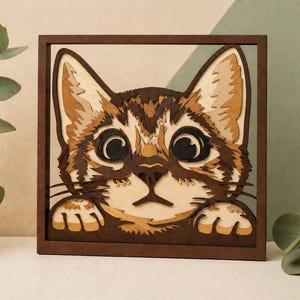 Peut inclure: Une œuvre d'art en bois multicouche représentant un visage de chat. Le chat a de grands yeux expressifs et un pelage brun et beige. L'œuvre est encadrée dans un carré marron foncé.