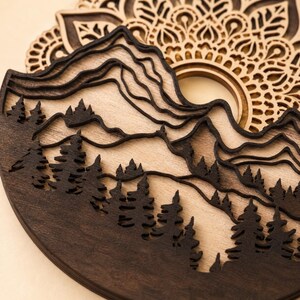 May include: A multi-layered wooden wall art piece featuring a dark brown laser-cut silhouette of a mountain range and a dense pine forest. Behind the mountains, a light-colored wooden circular mandala pattern with intricate geometric cutouts.