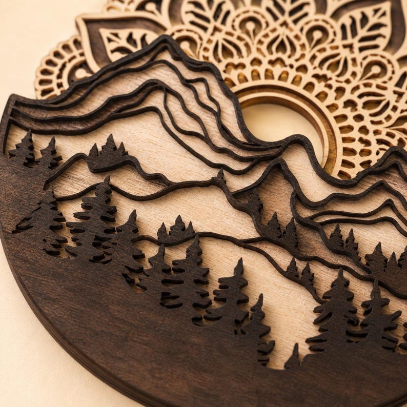 May include: A multi-layered wooden wall art piece featuring a dark brown laser-cut silhouette of a mountain range and a dense pine forest. Behind the mountains, a light-colored wooden circular mandala pattern with intricate geometric cutouts.