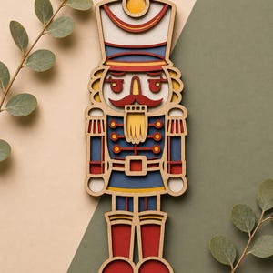 May include: A wooden, layered nutcracker figurine in a classic soldier design. The figurine features a white hat with red and yellow accents, a blue jacket with red details, and red boots. The background is a split of beige and green.