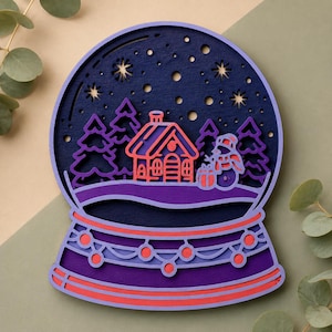 May include: A layered wooden snow globe decoration with a winter scene. The design features a house, snowman, and trees in shades of purple, red, and blue. The base has a decorative red and purple trim. The background is a dark blue with star cutouts.