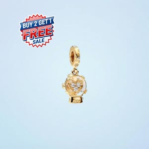 May include: A gold-colored charm with a snow globe design. The charm features a heart-shaped design with clear crystals inside the globe. The charm has a loop at the top for attaching to a bracelet or necklace. The image also includes a "BUY 2 GET 1 FREE SALE" sticker.