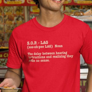 Può includere: Una maglietta rossa con testo bianco. Il testo recita: "S.O.P. - LAG (ess-oh-pee LAG) Noun. Il ritardo tra l'ascolto delle istruzioni e la realizzazione che non hanno senso." La maglietta è indossata.