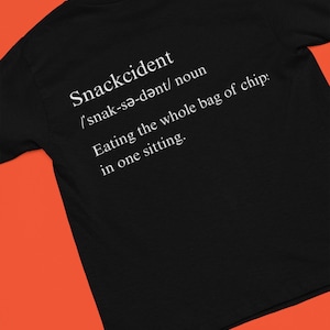 May include: Black t-shirt with the definition of "Snackcident" printed in white. The definition includes the pronunciation, and the phrase "Eating the whole bag of chips in one sitting."