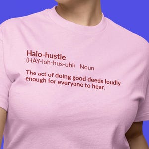May include: A light pink t-shirt with the text "Halo-hustle" in red, along with a definition. The definition reads: "The act of doing good deeds loudly enough for everyone to hear."