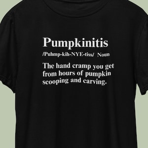 Pode incluir: Camiseta preta com a palavra "Pumpkinitis" em branco, juntamente com uma definição de cãibra na mão por esculpir abóboras. A camiseta é exibida em um cabide de madeira.