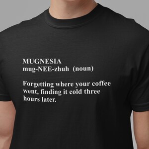 Pode incluir: Camiseta preta com a palavra "MUGNESIA" e a definição: "Esquecer para onde foi o seu café, encontrá-lo frio três horas depois."