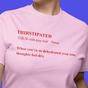 May include: A light pink t-shirt with the word "THIRSTIPATED" in red, along with its pronunciation and definition. The shirt also features the phrase "When you're so dehydrated even your thoughts feel dry."