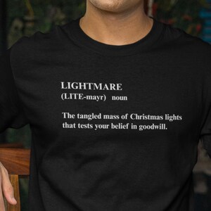 Pode incluir: Camiseta preta com a palavra "LIGHTMARE" e sua pronúncia entre parênteses. Abaixo, o texto define "LIGHTMARE" como "A massa emaranhada de luzes de Natal que testa sua crença na boa vontade."