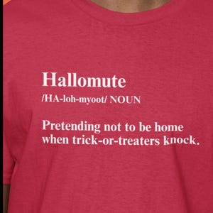 Pode incluir: Camiseta vermelha com a palavra "Hallomute" em branco, sua pronúncia e definição: "Fingir que não está em casa quando os pedintes de doces batem." Uma peça de roupa divertida com tema de Halloween.