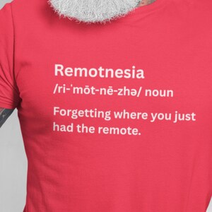 May include: Red t-shirt with the word "Remotnesia" and its pronunciation in white text. The definition, "Forgetting where you just had the remote," is also printed on the shirt. The shirt is a solid red color.