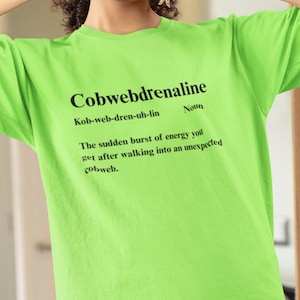 Pode incluir: Camiseta verde neon com a palavra "Cobwebdrenaline" em preto e uma definição. A definição descreve a explosão repentina de energia após entrar em uma teia de aranha inesperada.