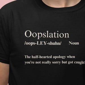May include: Black t-shirt with the word "Oopslation" in white, along with its pronunciation and definition. The definition reads: "The half-hearted apology when you're not really sorry but got caught."