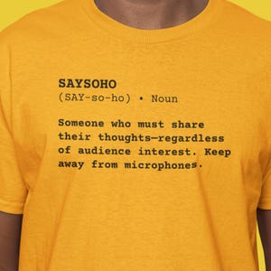 Pode incluir: Camiseta amarelo mostarda com a definição de "SAYSOHO" impressa em preto. A definição inclui a pronúncia, a classe gramatical e uma descrição humorística. A camiseta é de gola redonda.