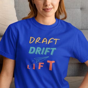 Può includere: T-shirt blu reale con le parole "DRAFT DRIFT LIFT" in arancione, verde acqua e giallo. Una grafica di aeroplano di carta è tra "DRIFT" e "LIFT". La maglietta è a girocollo.