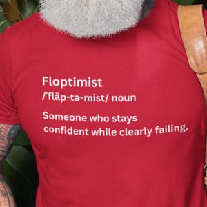 May include: Red t-shirt with the word "Floptimist" and its phonetic pronunciation, followed by the definition: "Someone who stays confident while clearly failing."