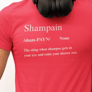 May include: Red t-shirt with the word "Shampain" in white, along with a definition. The definition describes the stinging sensation when shampoo enters the eye. Black headphones rest on the shoulders.