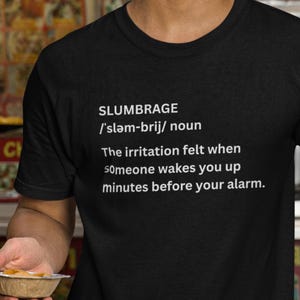 May include: Black crew neck t-shirt with the word "SLUMBRAGE" and its definition printed in white. The definition reads: "The irritation felt when someone wakes you up minutes before your alarm."