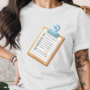 May include: A light-colored t-shirt featuring a clipboard graphic. The clipboard has a wooden frame, a blue clip, and a checklist with checkmarks. The design is simple and clean, suitable for various themes.