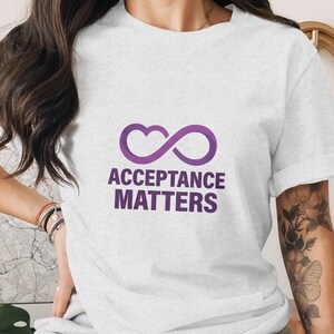 May include: A light gray t-shirt with a purple infinity heart symbol and the words "ACCEPTANCE MATTERS" in purple. The shirt is made of a soft material and has short sleeves.