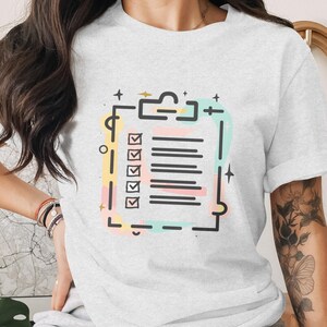 May include: A light gray t-shirt featuring a checklist design. The checklist is outlined in black with colorful accents of yellow, pink, and teal. The design includes checkmarks next to the list items.