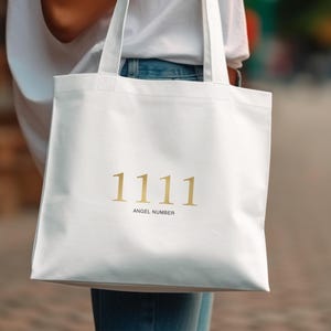 May include: A white tote bag featuring the gold text "1111 ANGEL NUMBER." The bag is carried by someone wearing a white top and blue jeans. The background is blurred, implying an outdoor location.