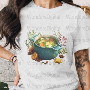 May include: A light gray t-shirt featuring a watercolor illustration of a teal pot filled with soup, surrounded by herbs, spices, and sliced lemons. The text "Wonders Digital" is printed on the shirt.
