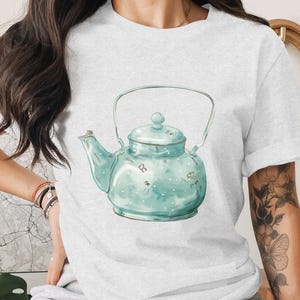 May include: Light gray t-shirt featuring a watercolor illustration of a teal teapot with white polka dots. The teapot has a curved handle and a lid with a small knob. The design is centered on the shirt.