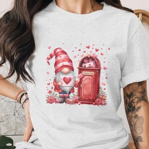 May include: A light gray t-shirt featuring a Valentine's Day gnome holding a heart-shaped card. The gnome stands next to a red mailbox labeled "Valentine Shop", surrounded by hearts and roses. The design is a watercolor illustration.