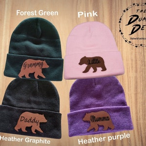 May include: Four knit beanies in different colors with brown bear patches. The beanies are forest green, pink, heather graphite, and heather purple. The bear patches have the words "Grammy", "Little", "Daddy", and "Momma" on them.