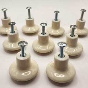 May include: A collection of ten off-white ceramic cabinet knobs with silver-colored screws. The knobs have a rounded base and a cylindrical stem, creating a classic and functional design for furniture or cabinetry.