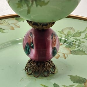 Puede incluir: Un plato decorativo de varios niveles con una base verde claro y una parte superior a juego. El plato presenta una pieza central esf&eacute;rica de color rosa oscuro con detalles pintados y adornos met&aacute;licos de color bronce. Los patrones florales adornan la base.