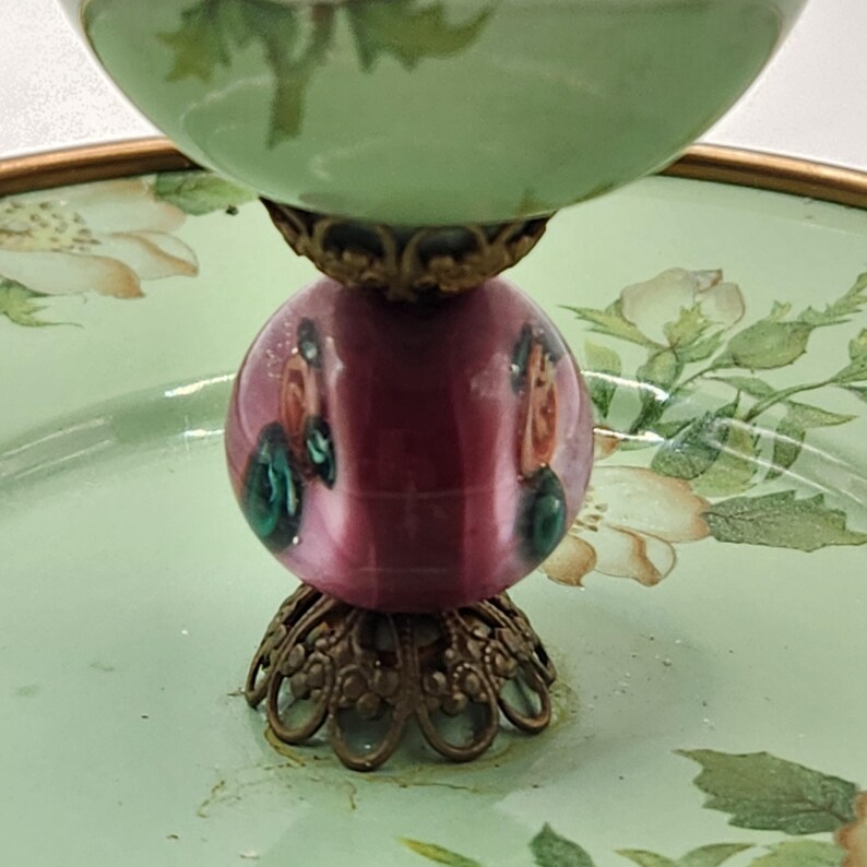 Puede incluir: Un plato decorativo de varios niveles con una base verde claro y una parte superior a juego. El plato presenta una pieza central esf&eacute;rica de color rosa oscuro con detalles pintados y adornos met&aacute;licos de color bronce. Los patrones florales adornan la base.