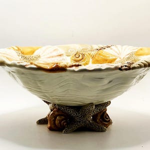 May include: A decorative ceramic bowl with a seashell design. The bowl is white with a wavy texture and features various seashells in shades of tan and brown around the rim. The base is supported by starfish and shell accents.