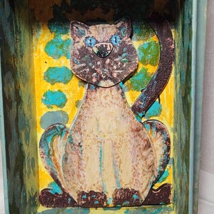 May include: A wooden cat figurine with a light brown, beige, and gray painted finish. The cat is sitting with its tail curled around its body. The background is a yellow and blue painted surface.