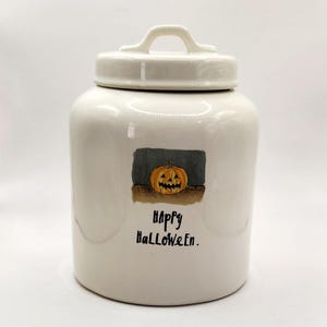 May include: White ceramic canister with a lid and a handle. The canister has a black and gray illustration of a jack-o'-lantern with the text "Happy Halloween."