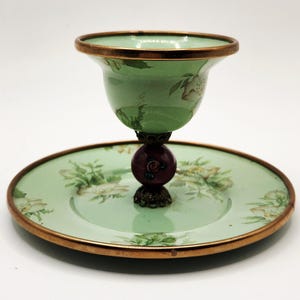 Puede incluir: Un plato decorativo vintage verde y dorado con una taza a juego. La taza y el plato tienen un dise&ntilde;o floral. La taza est&aacute; sostenida por una base de color rojo oscuro y bronce. El borde de la taza y el plato est&aacute; adornado con oro.