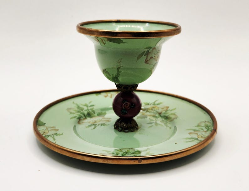 Puede incluir: Un plato decorativo vintage verde y dorado con una taza a juego. La taza y el plato tienen un dise&ntilde;o floral. La taza est&aacute; sostenida por una base de color rojo oscuro y bronce. El borde de la taza y el plato est&aacute; adornado con oro.