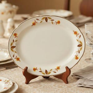 May include: An oval-shaped cream-colored ceramic serving platter featuring a delicate border pattern of orange and yellow flowers with brown leaves. The platter is displayed on a dark wooden stand, showcasing its smooth finish and gold-toned rim.