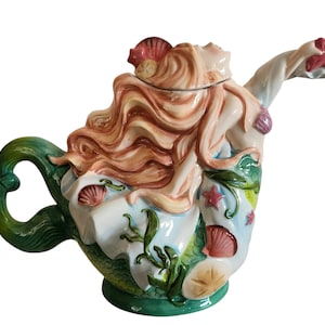 Puede incluir: Una tetera de cerámica con forma de sirena. La tetera presenta una sirena con cabello castaño rojizo, un top blanco y una cola verde. La tetera está decorada con conchas marinas, estrellas de mar y algas marinas. El asa es verde y el pico termina con una estrella de mar.