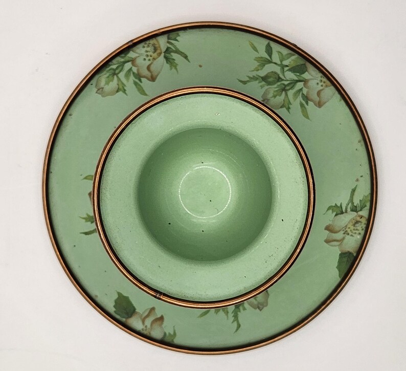 Puede incluir: Un juego de platos de cer&aacute;mica verde claro con un dise&ntilde;o floral. El juego incluye un cuenco peque&ntilde;o y centrado y un plato m&aacute;s grande, ambos con un borde color cobre. El plato presenta un patr&oacute;n de flores blancas y beige con hojas verdes.